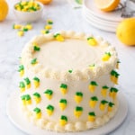 Triple Lemon Layer Cake decorated with tiny royal icing lemons, on a white cake plate with marble surface and lemons in the background.