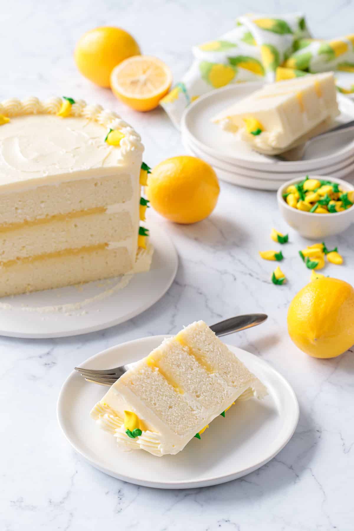 Triple Lemon Layer Cake, slice on a white plate in the foreground with the whole cake and another slice in the background along with some fresh Meyer lemons.