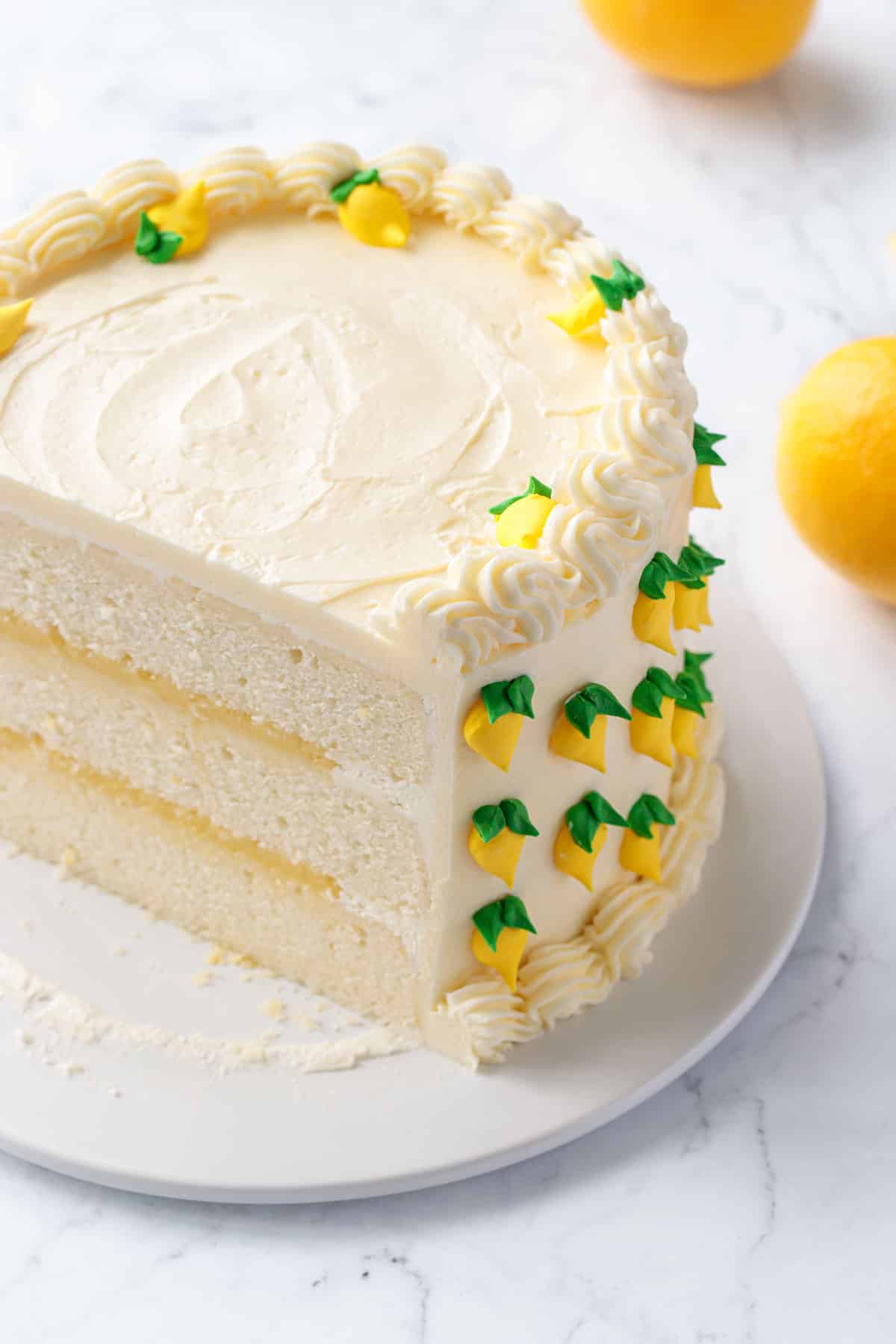 Triple Lemon Layer Cake on a white cake plate, front sliced off to show the layers of cake, curd, and frosting inside.