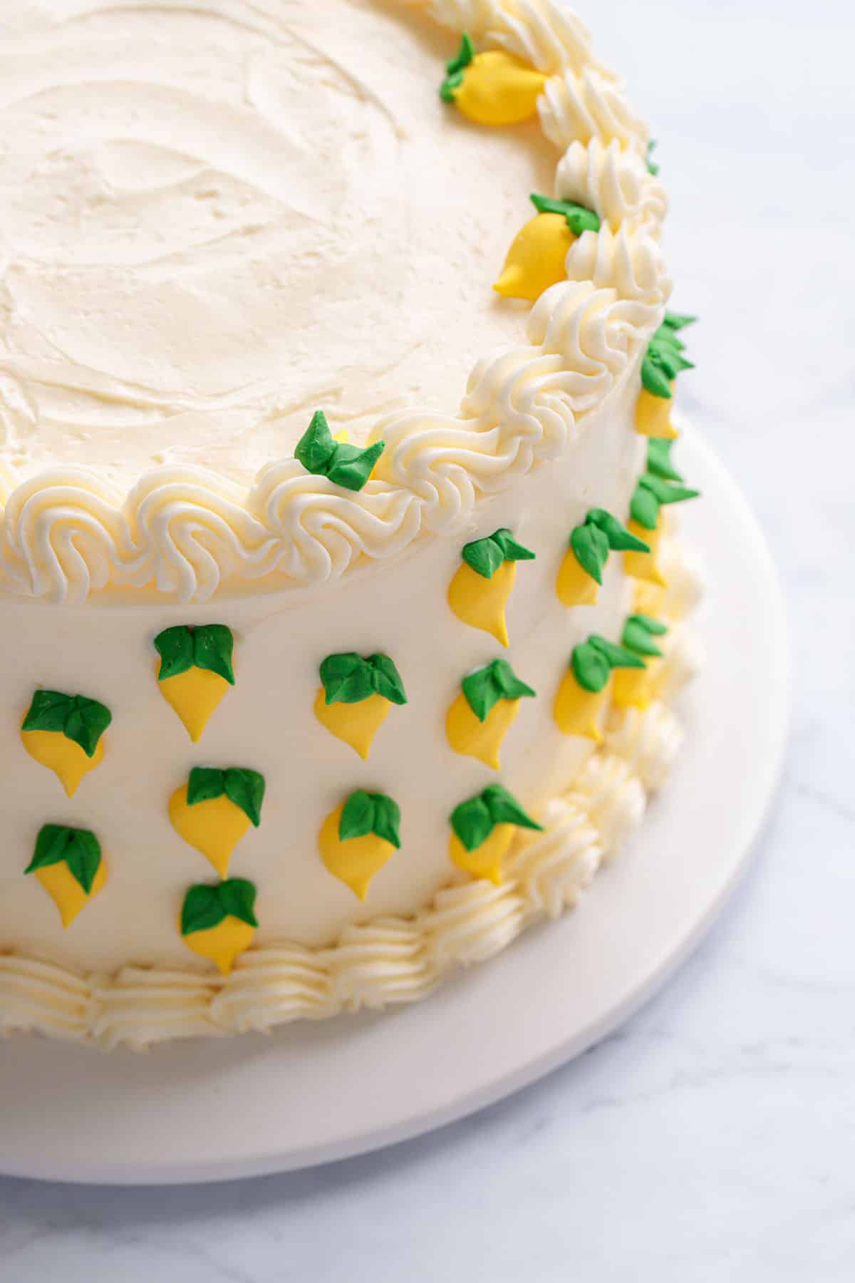 Triple Lemon Layer Cake decorated with white frosting, piped borders, and tiny royal icing lemons with leaves all around the outside.