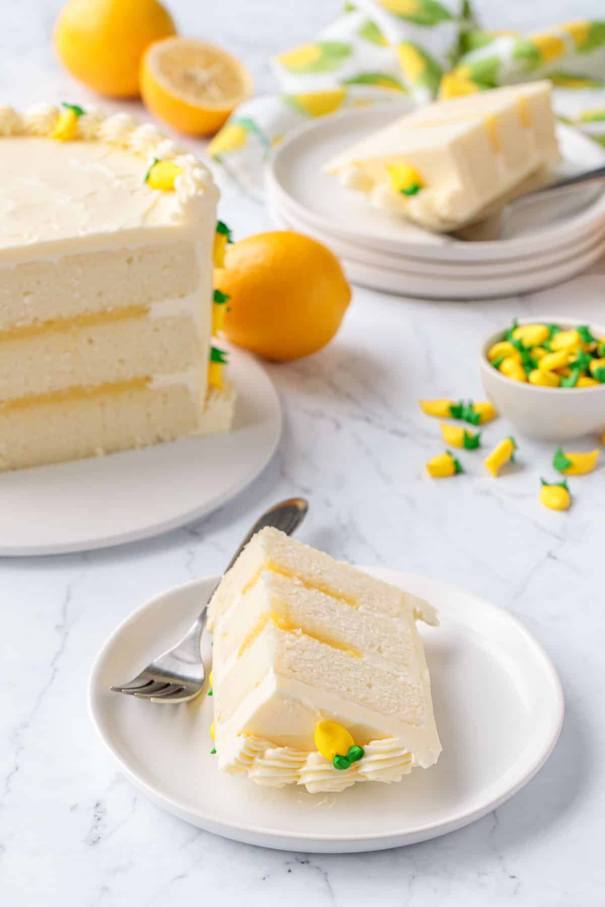 Triple Lemon Layer Cake, slice on a white plate in the foreground with the whole cake and another slice in the background along with some fresh Meyer lemons.