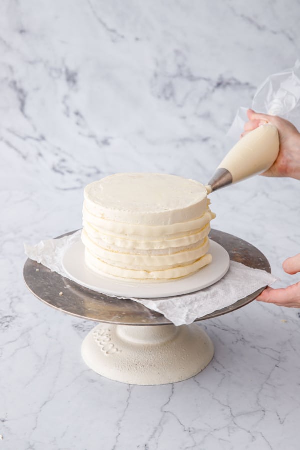 After chilling the crumb coat, piping a thicker final layer of frosting onto the sides and top of cake.