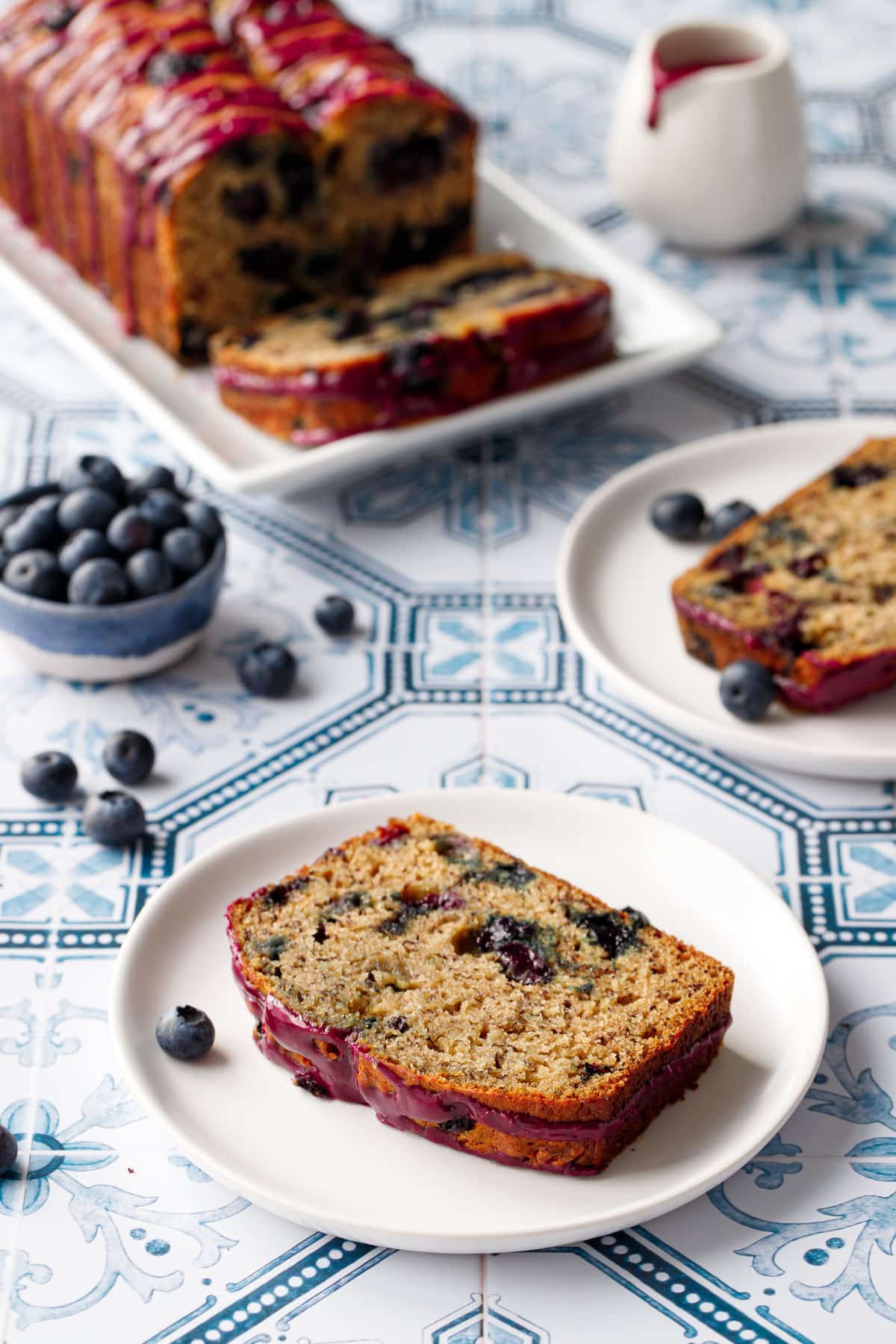 Two plates with slices of Blueberry Banana Bread on a blue and white tile background, with bowl of blueberries and the remainder of the loaf in the background.