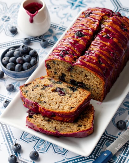 Loaf of Blueberry Banana Bread on a white rectangular plate with two slices lying down on a blue and white tile background.