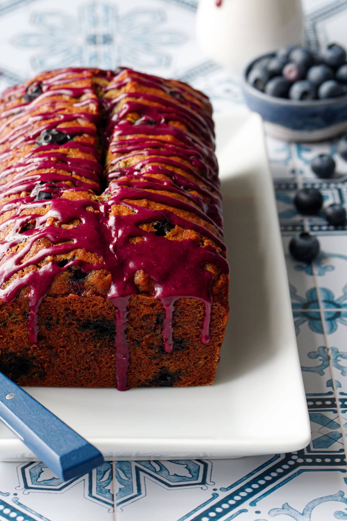 Loaf of Blueberry Banana Bread on a white plate, close up to show the drips of the magenta-colored blueberry glaze down the side.