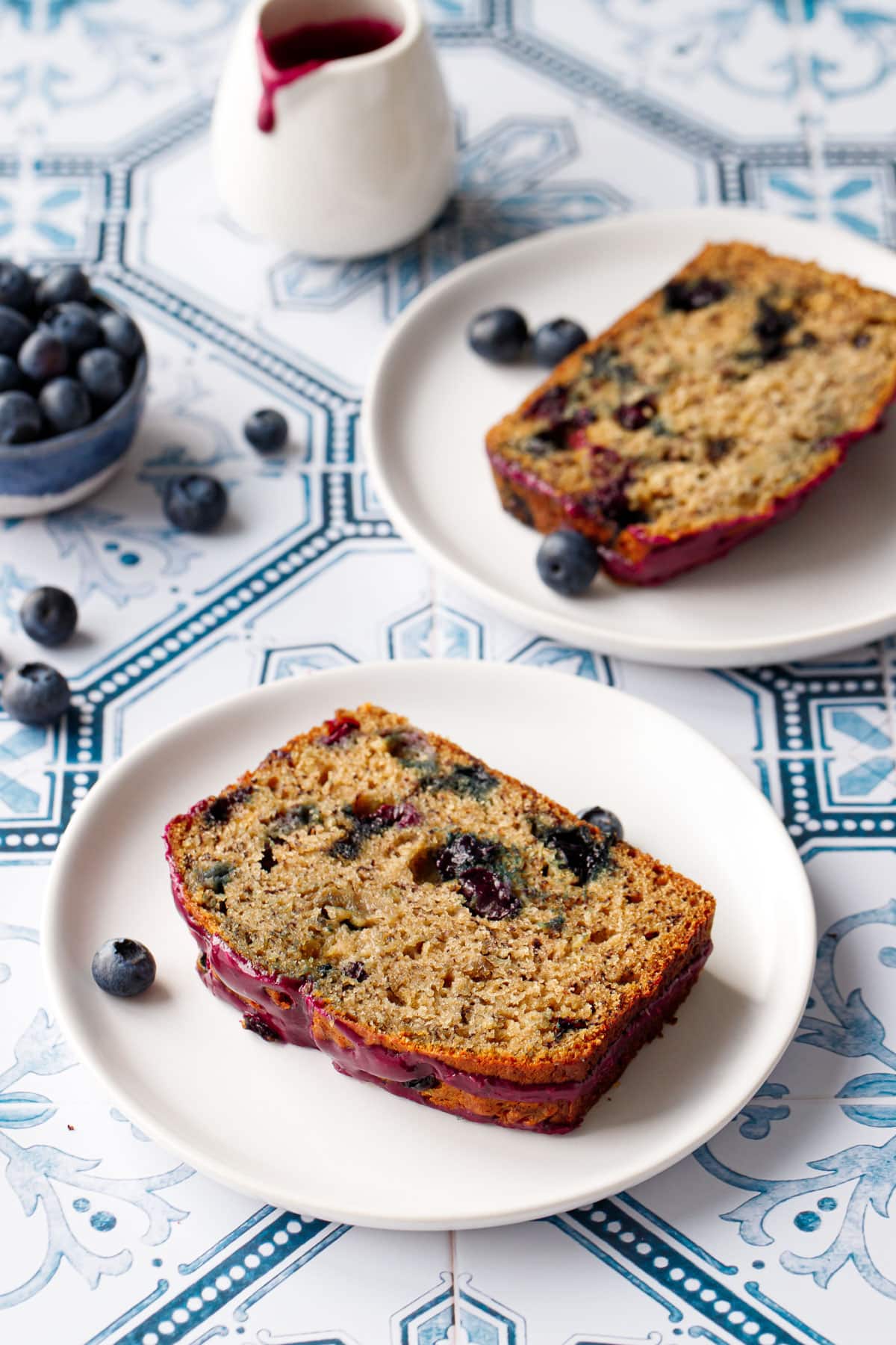 Two plates with slices of Blueberry Banana Bread on a blue and white tile background, with bowl of blueberries and pouring vessel of glaze with a drip on the side.