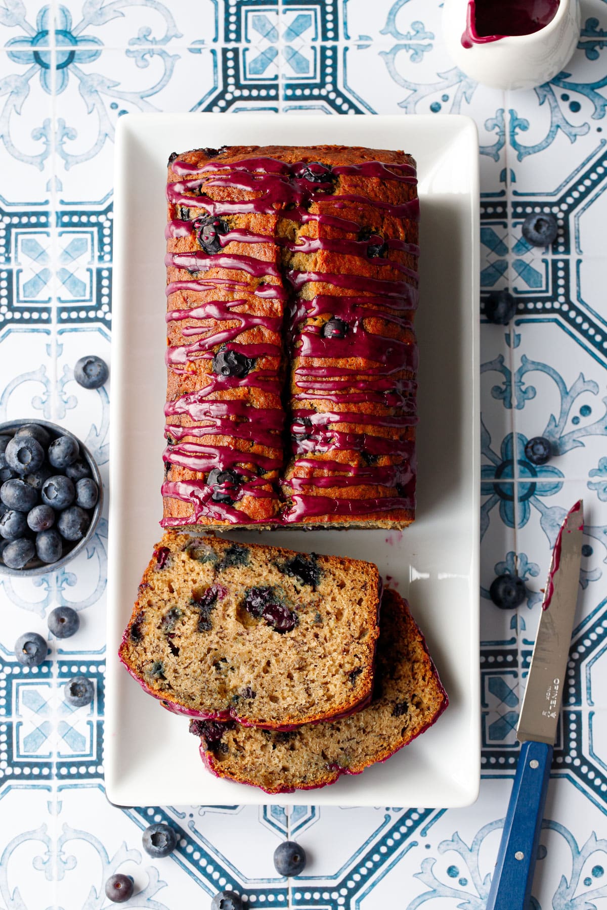 Overhead, loaf of Blueberry Banana Bread on a white rectangular plate with two slices lying down on a blue and white tile background with blue knife and small bowl of blueberries and extra glaze.