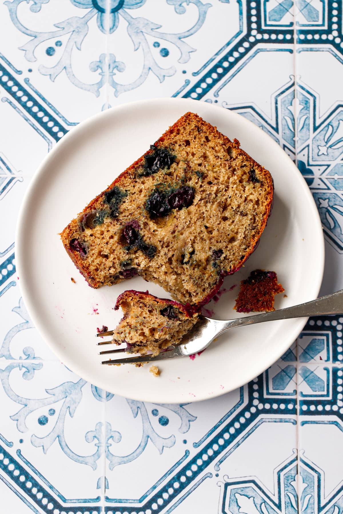 Closeup, single slice of Blueberry Banana Bread on a white plate with a forkful taken out of it to show the moist texture.