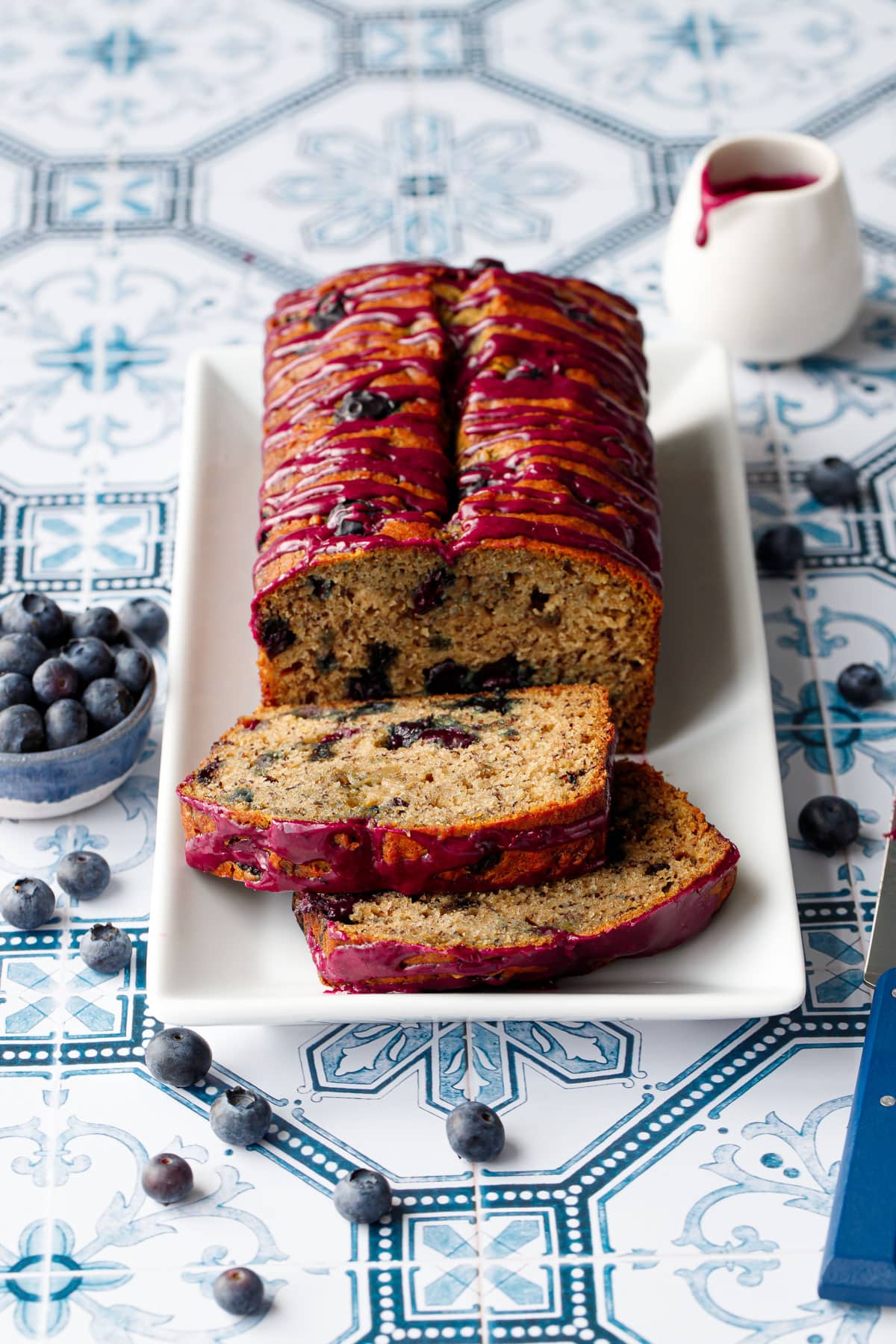 Loaf of Blueberry Banana Bread on a white rectangular plate with two slices lying down on a blue and white tile background.