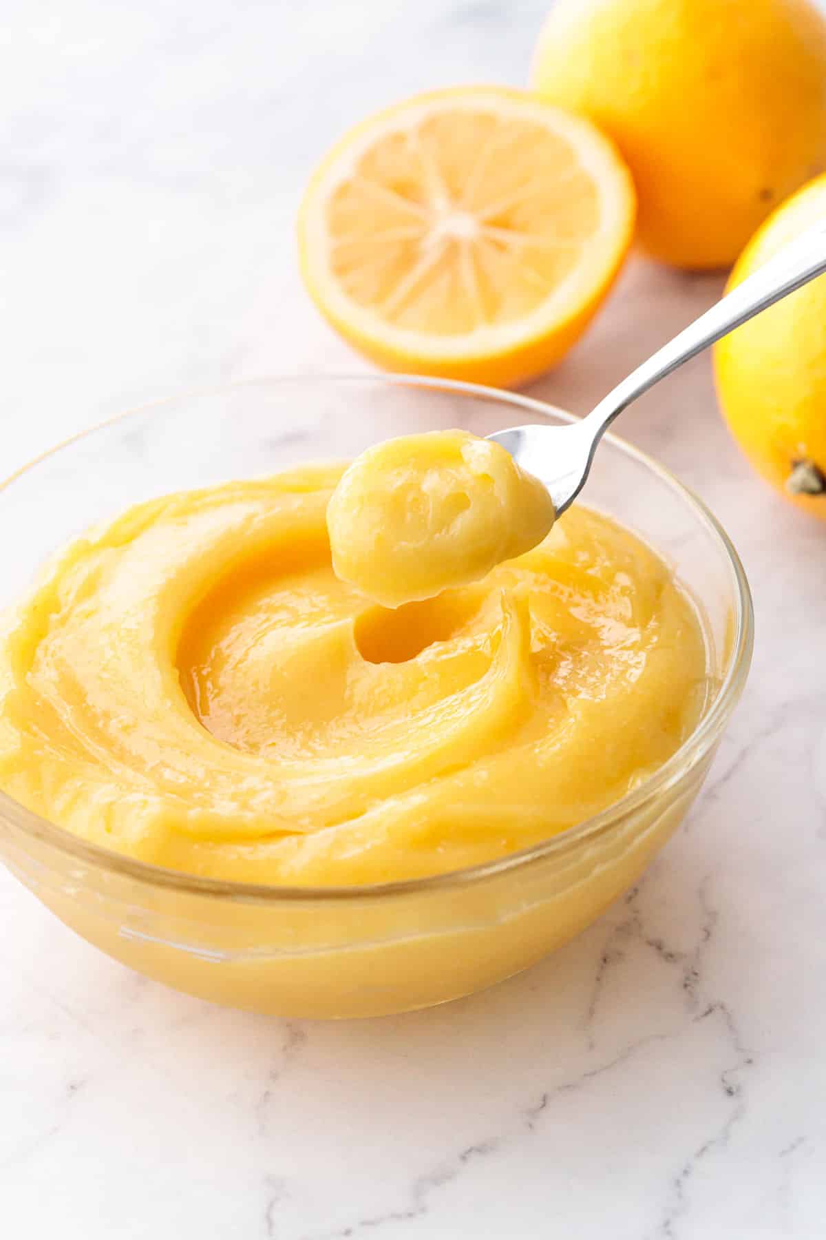 Spoonful of Meyer Lemon Curd being lifted out of a glass bowl, with fresh lemons in the background.