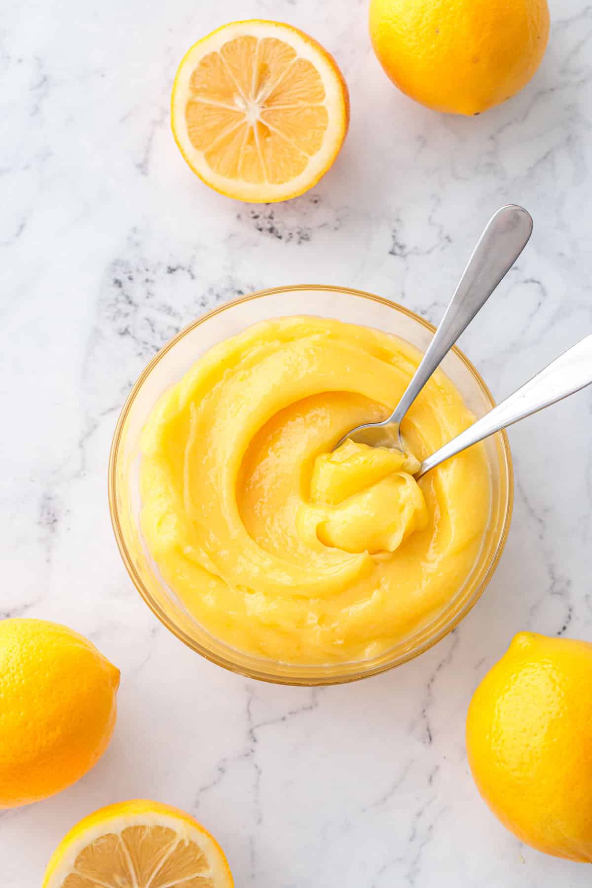 Overhead, bowl of Meyer Lemon Curd with two spoons, with fresh whole and halved lemons scattered around on the marble surface.