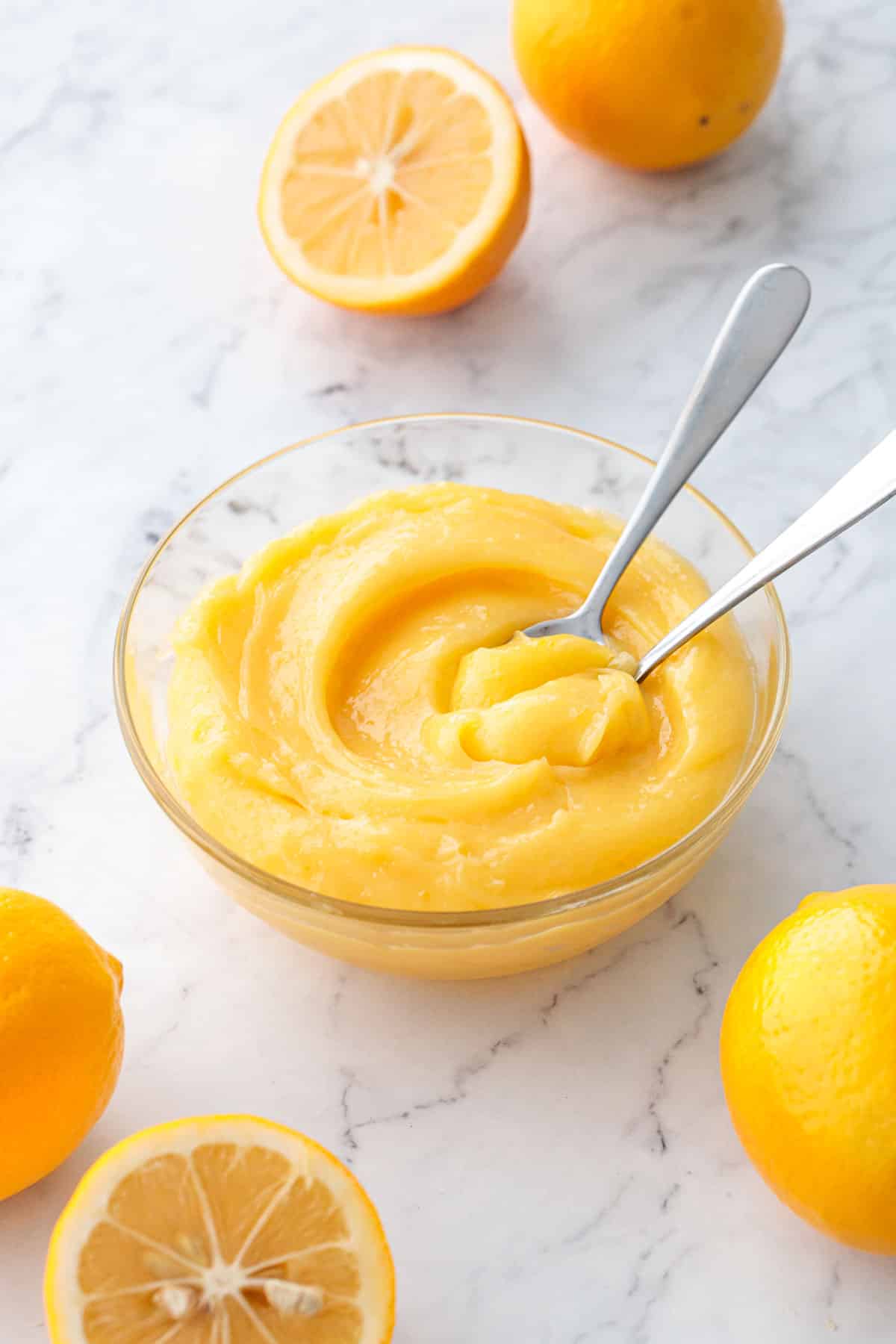 Glass bowl filled with a swirl of Meyer Lemon Curd and two spoons, with fresh lemons scattered around.