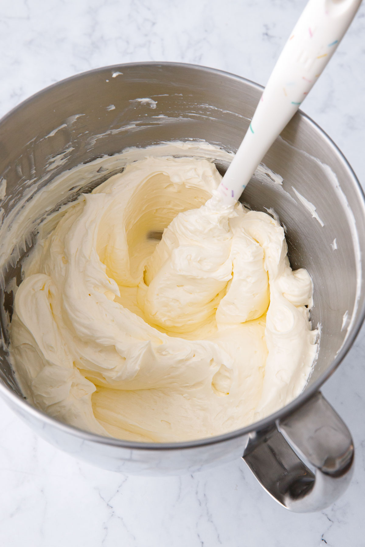 Bowl of Swiss Meringue Buttercream in a metal mixing bowl with white sprinkle spatula.