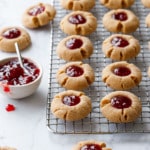 Rows of Peanut Butter & Jam Thumbprints on a wire baking rack, with bowl of strawberry jam and a few more cookies scattered around it.