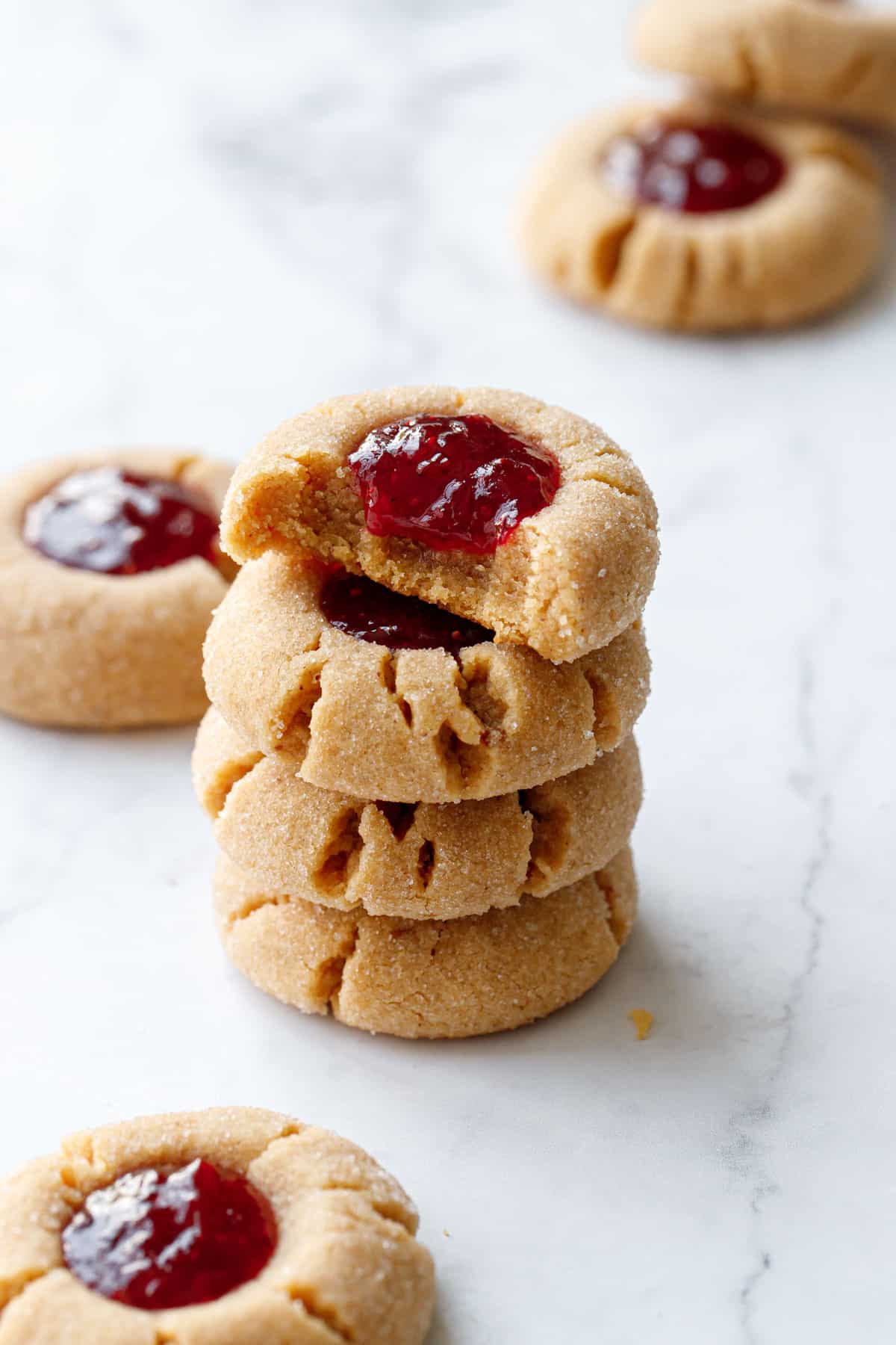 Stack of Peanut Butter & Jam Thumbprints, top cookie with a bite taken out to show the consistency, on a marble background.