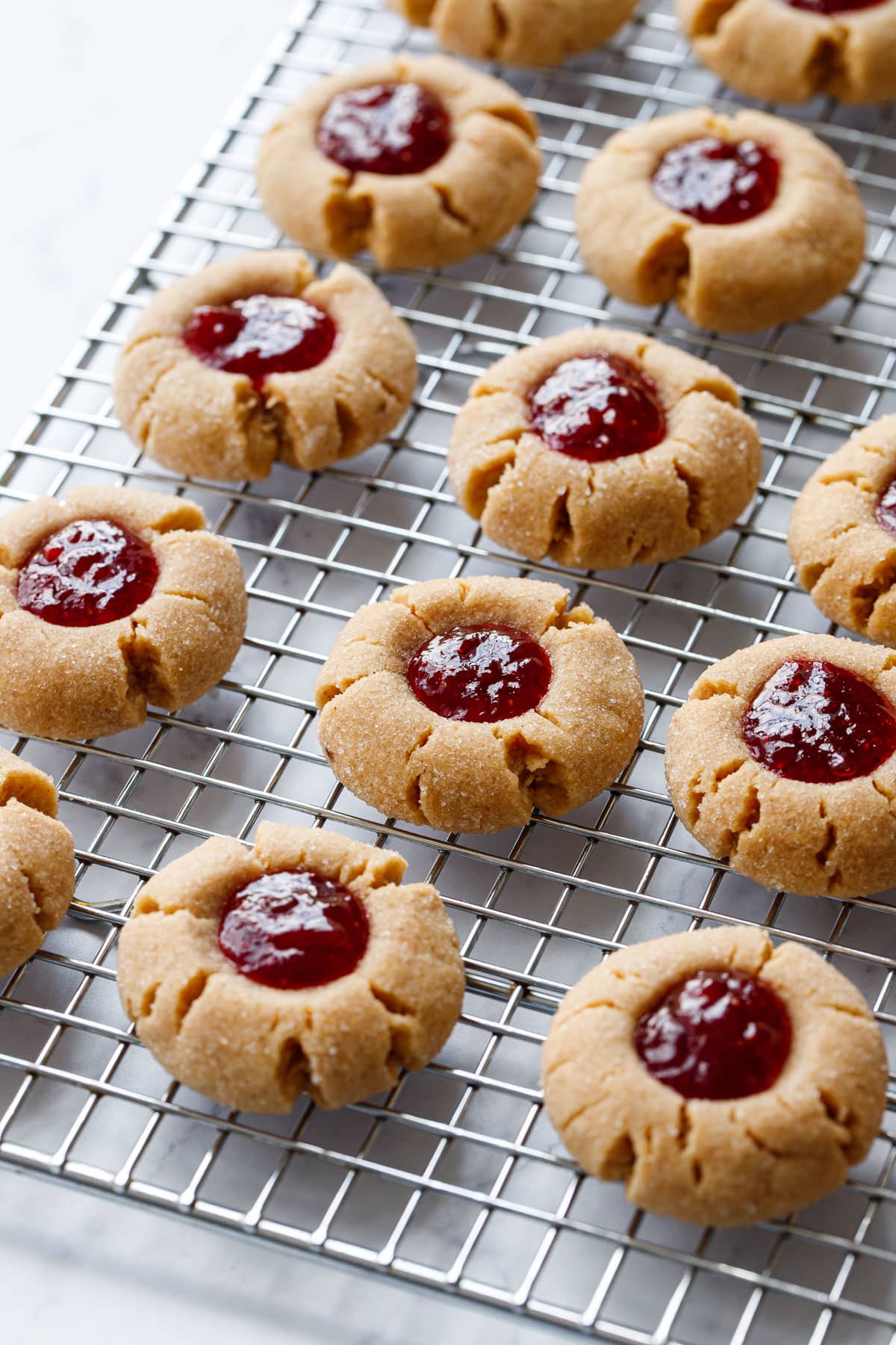 Wire baking rack with rows of Peanut Butter & Jam Thumbprints filled with shiny strawberry jam on a marble background.