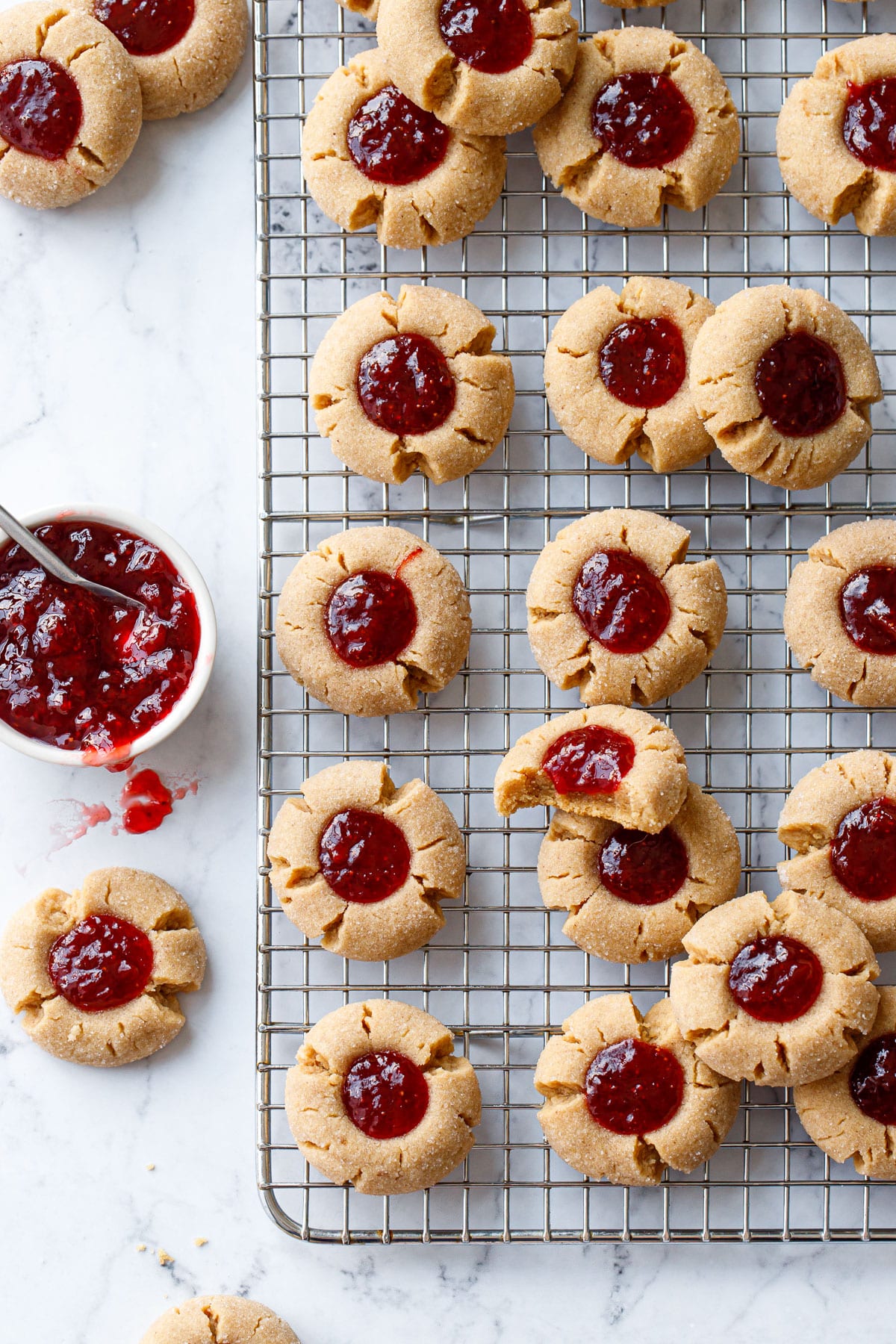 Overhead, Peanut Butter & Jam Thumbprint cookies on a wire baking rack, one bitten cookie and a bowl of strawberry jam on the side.