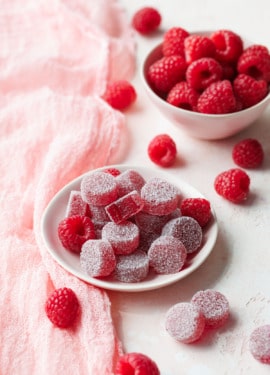 Small dish of Homemade Raspberry Pectin Gummies on a light pink background with linen and bowl of raspberries in the background.