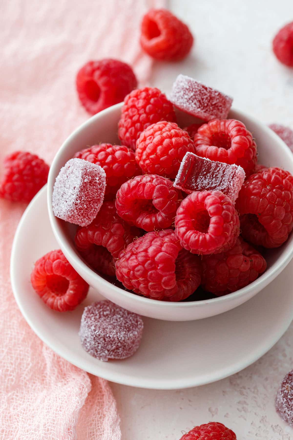 Bowl of fresh raspberries with a few Raspberry Gummy candies sitting on top and scattered around, with a light pink background and linen.
