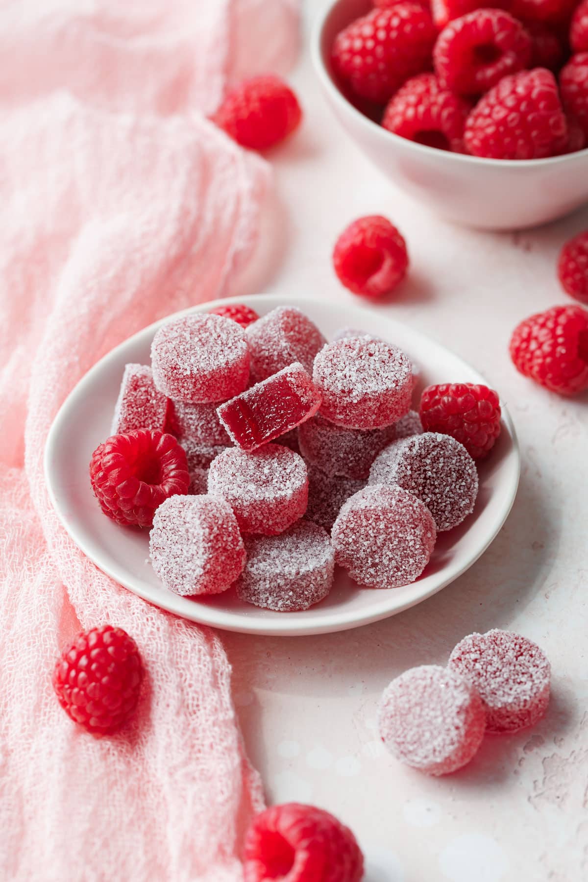 Small dish of Homemade Raspberry Pectin Gummies on a light pink background with linen and bowl of raspberries in the background.