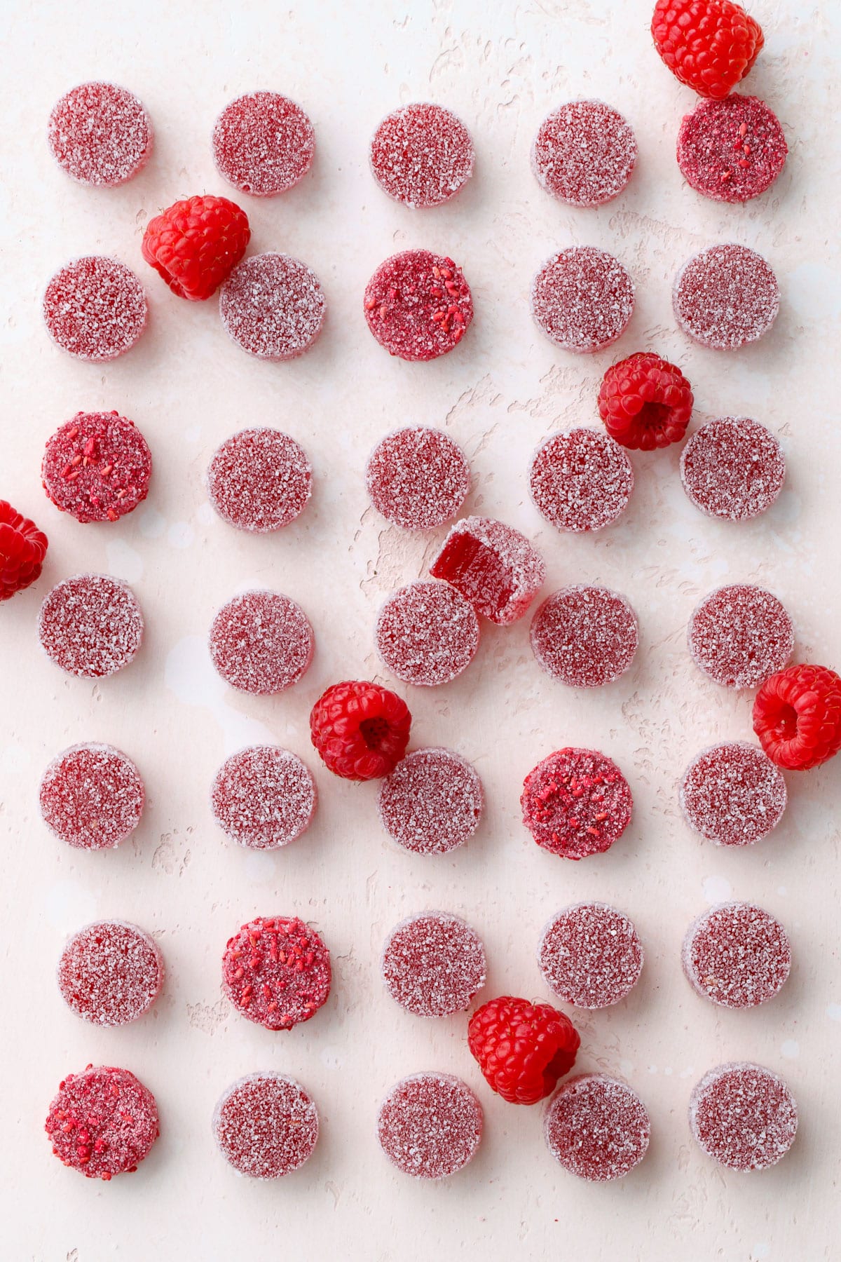 Overhead, neat rows of Homemade Raspberry Pectin Gummies with one bitten candy and a few raspberries breaking up the grid.