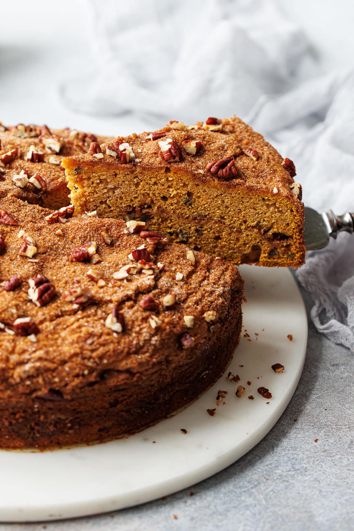 Cake server lifting a slice of Pumpkin Pecan Coffee Cake on a marble cake plate with a gray background.