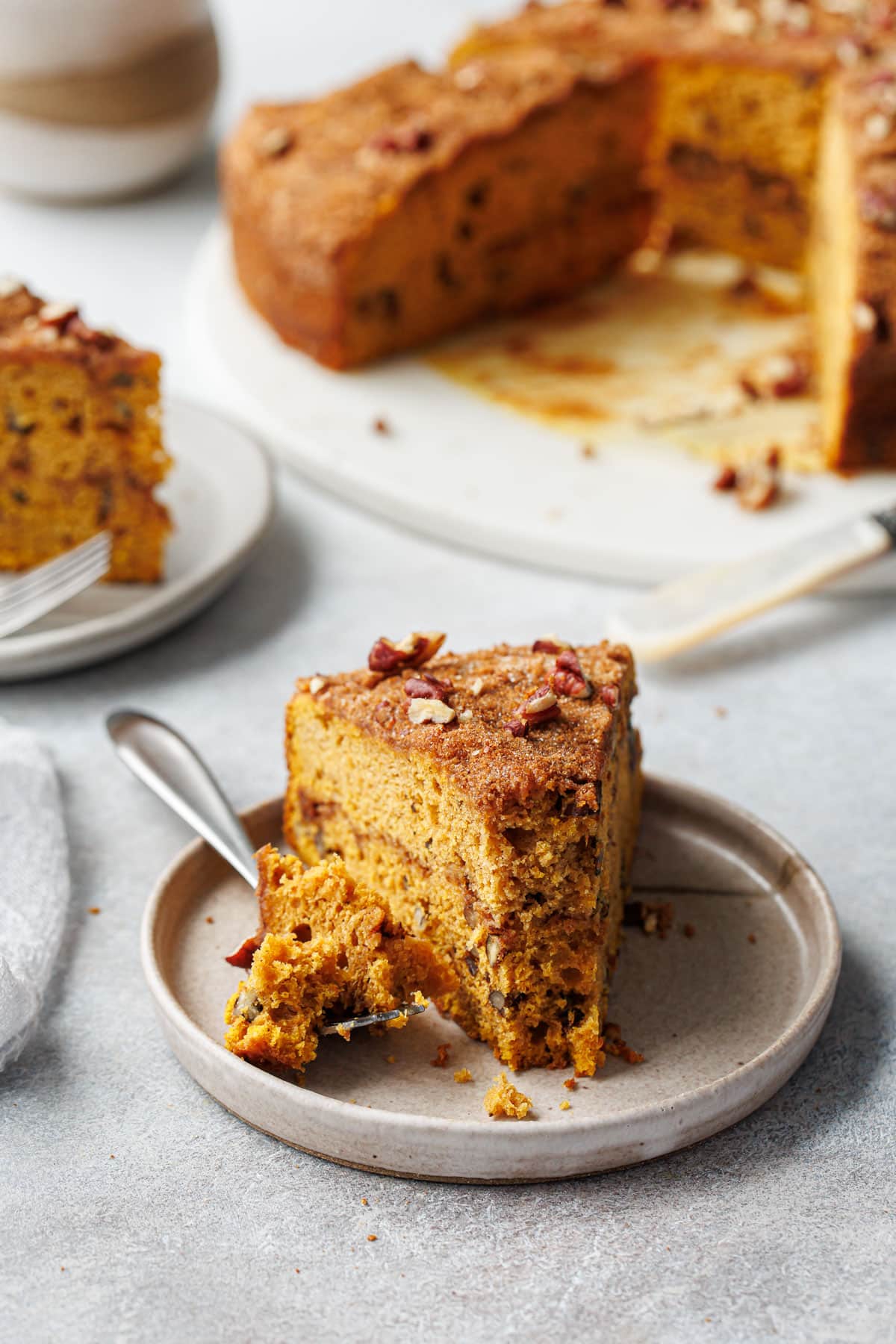 Slice of Pumpkin Pecan Coffee Cake with a forkful taken off the end to show the moist texture, rest of the cake and coffee cup out of focus in the background.