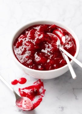 White bowl filled with Classic Cranberry Sauce and two spoons, plus another spoon with a smear of cranberry sauce on the marble surface.