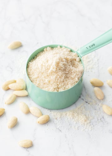 Mint green measuring cup overflowing with Homemade Almond Flour, on a marble background with a bit of flour and whole blanched almonds scattered around.