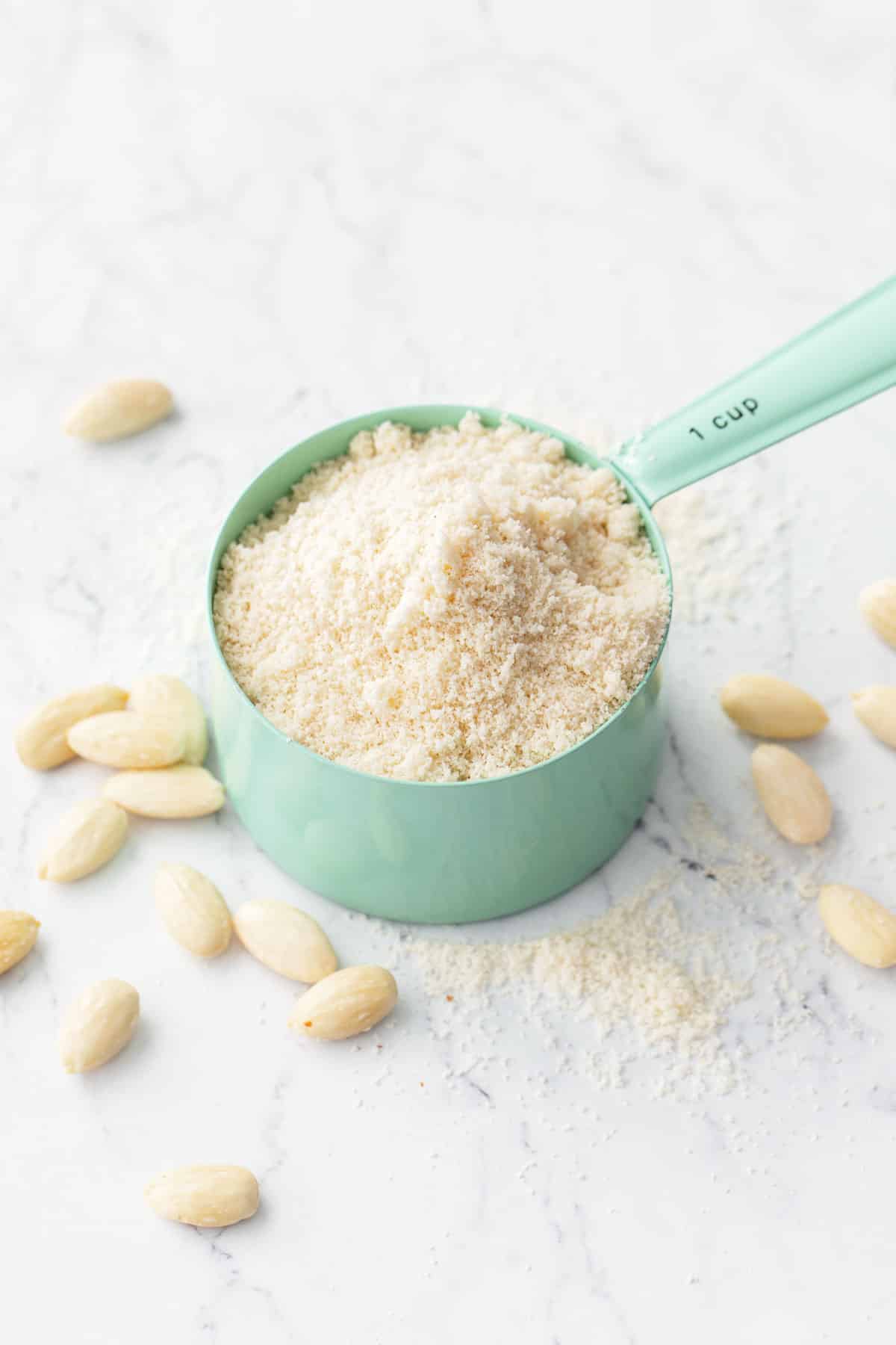 Mint green measuring cup overflowing with Homemade Almond Flour, on a marble background with a bit of flour and whole blanched almonds scattered around.