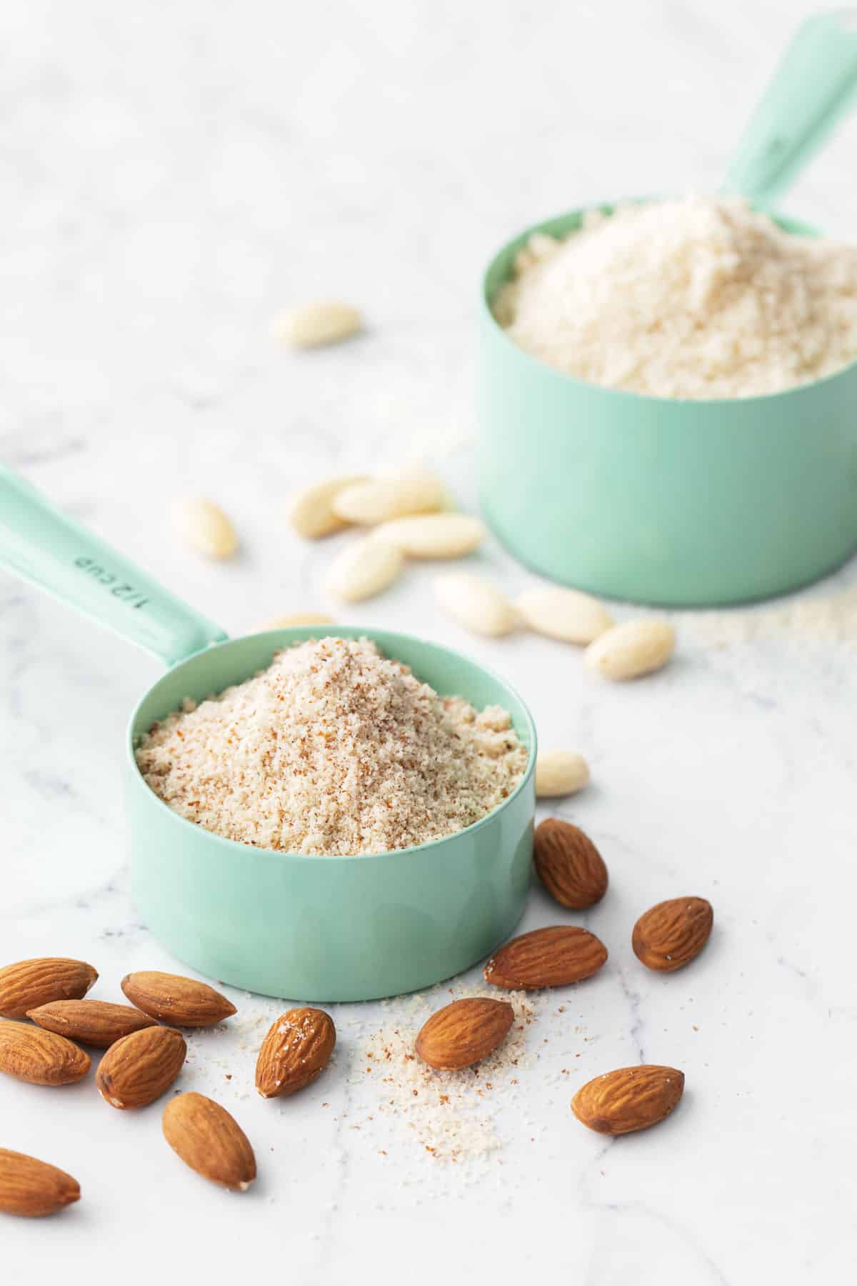 Two measuring cups, one filled with homemade almond flour made from blanched/unpeeled almonds, the other made from raw almonds which is darker and speckly in appearance.