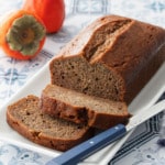 Loaf of Spiced Persimmon Bread on a white cake plate, two slices cut and laying down, with two hachiya persimmons in the background.