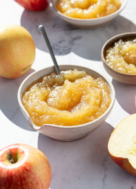 Bowl of homemade applesauce in direct sunlight, with a few smaller bowls and whole and halved apples around it.