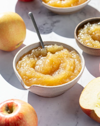 Bowl of homemade applesauce in direct sunlight, with a few smaller bowls and whole and halved apples around it.