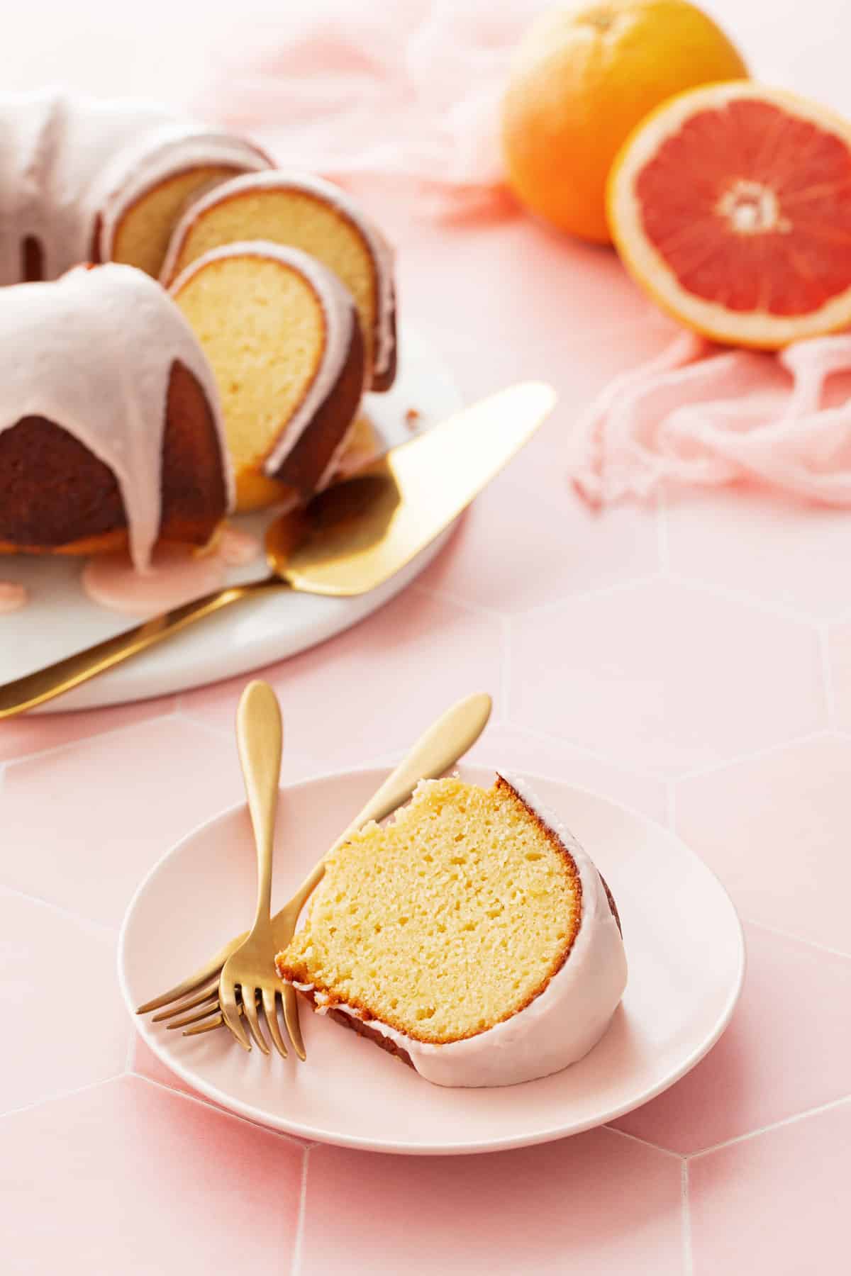Pink plate with a slice of Grapefruit Olive Oil Bundt Cake and two gold forks, the rest of the cake and a  cut grapefruit in the background.