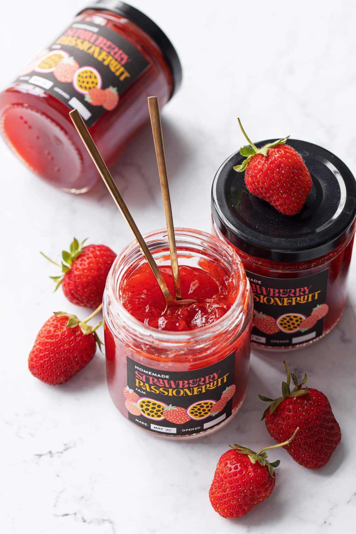 Three jars of homemade Strawberry Passionfruit Jam, one jar open with two gold spoons stuck inside, and a few fresh strawberries scattered around.