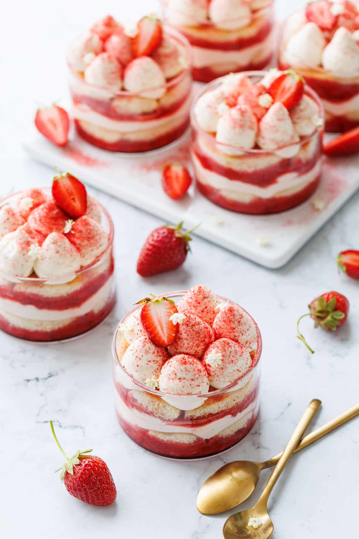 Strawberry Elderflower Tiramisu in individual clear ramekins, on a white marble background with two gold spoons and strawberries.