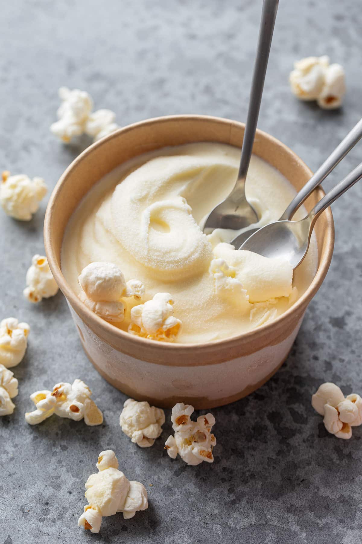 Brown paper pint container with a swirl of homemade Buttered Popcorn Ice Cream and three spoons scooping out bits, with a few scattered popcorn kernels.