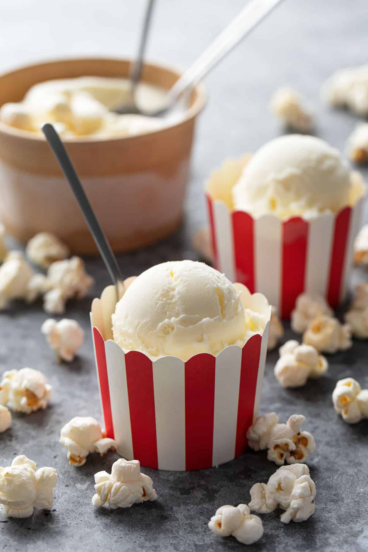 Red and white striped paper cups with scoops of Buttered Popcorn Ice Cream, with spoons and popped popcorn kernels scattered around.