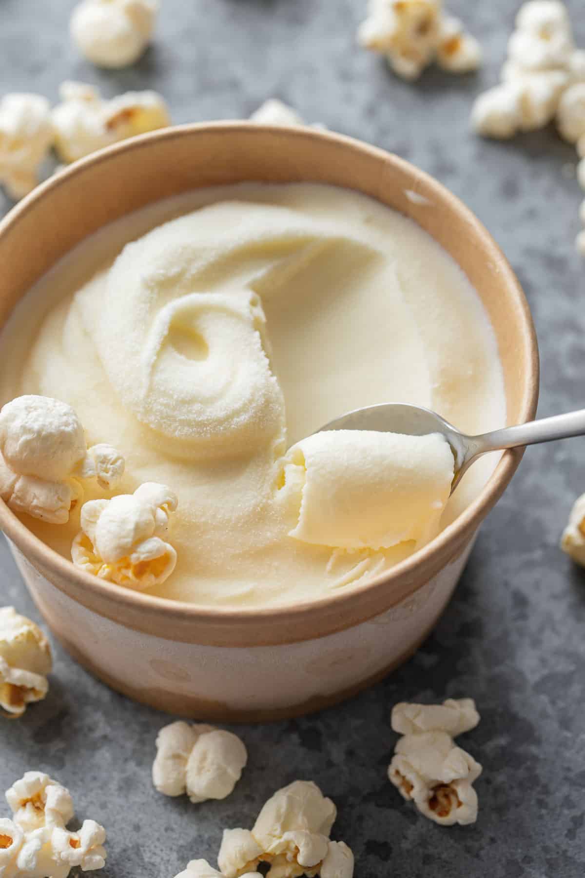 Closeup kraft pint container with Buttered Popcorn Ice Cream and a silver spoon scooping a swirl of ice cream, scattered popcorn kernels in the background.