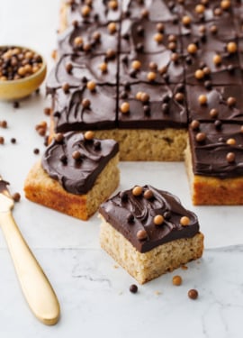 Cut pieces of Brown Butter Banana Cake with Chocolate Fudge Frosting on a marble surface with gold knife and topped with multi-color crispy pearls.
