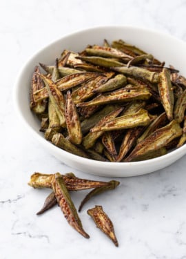 Bowl piled with Crispy Oven-Roasted Okra, a few laying on the side on a marble countertop.