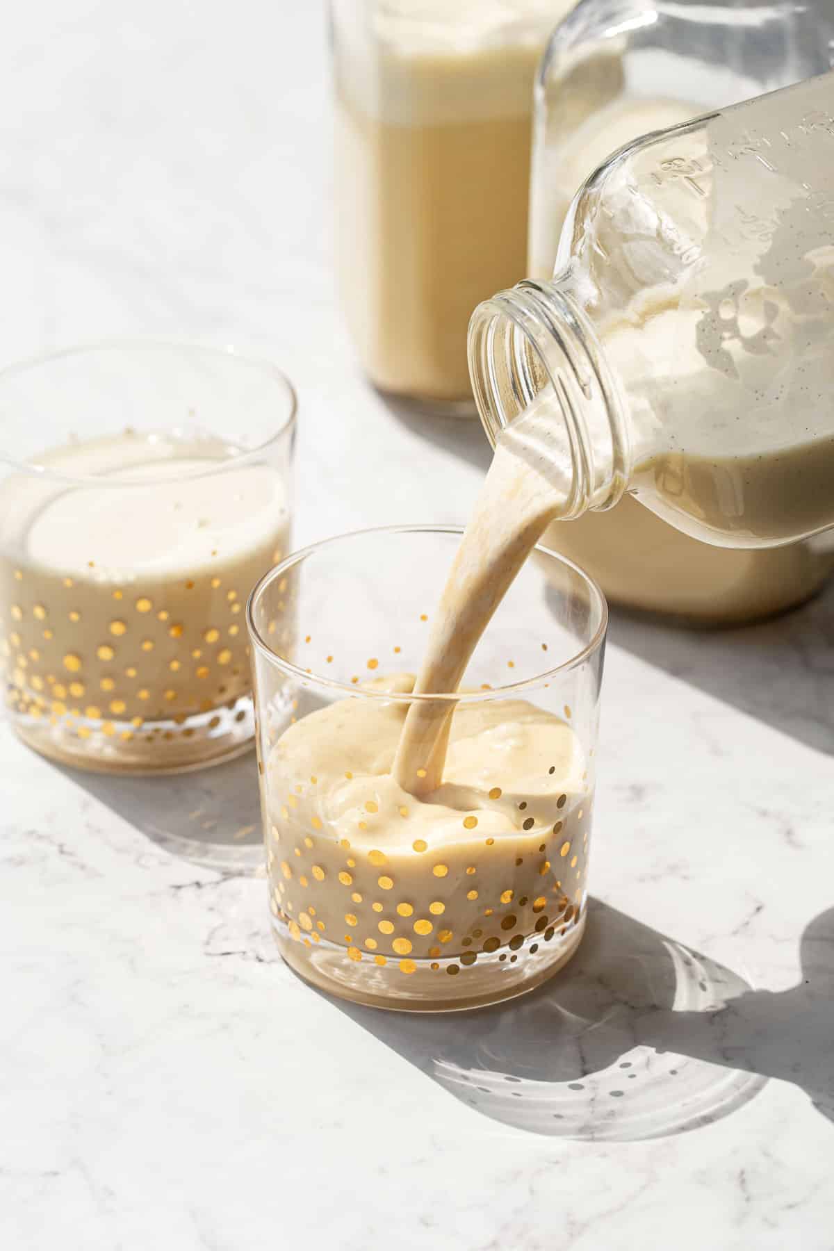 Pouring homemade aged eggnog into a gold speckled drinking glass with direct sunlight and strong shadows.