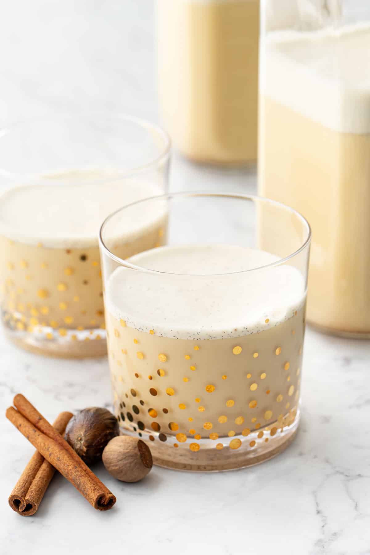 Gold speckled glasses filled with homemade eggnog, with whole cinnamon and nugmeg on the side and two bottles in the background.