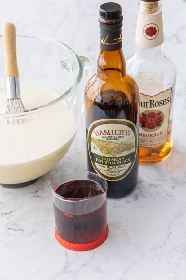 Bottles of dark rum and bourbon with a liquid measuring cup filled with rum, and bowl of eggnog mixture on the side.