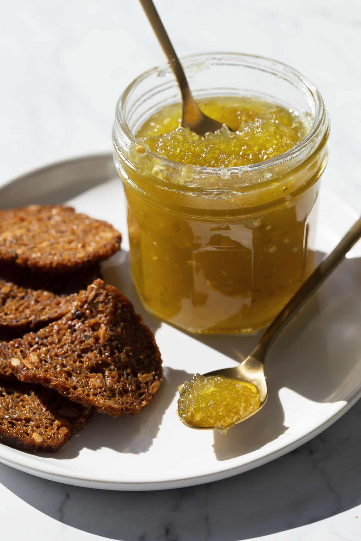Jar of Green Tomato Jam on a plate with dark rye crackers, with a spoonful of jam in the foreground to show the consistency.