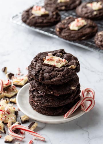 Stack of Chocolate Peppermint Bark Cookies on a small plate, with candy canes and broken up peppermint bark scattered around, rack of cookies cooling in the background.