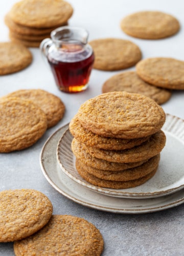 Stack of Chewy Maple Brown Butter Sugar Cookies on a ceramic plate, with a small glass pitcher of maple syrup and more cookies scattered around.