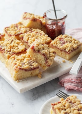 Squares of Sour Cream Crumb Cake with Jam Swirl and crumb topping on a marble serving plate with a jar of raspberry jam.