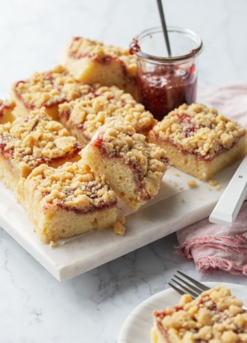 Squares of Sour Cream Crumb Cake with Jam Swirl and crumb topping on a marble serving plate with a jar of raspberry jam.